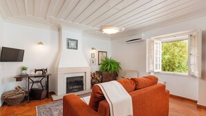 A living room with a fireplace, a sofa, a television, and a window with a view of greenery. - Casa da Avó Genoveva (Tomar)
