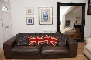 Un sofá de cuero marrón con un cojín con la bandera del Reino Unido, un cuadro enmarcado y un espejo en una habitación de paredes blancas.