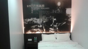 Habitación de hotel con cama, almohadas, mesita de noche con una botella y una pared con texto iluminado y una vista urbana.
