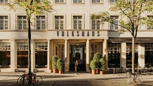 Un edificio con el letrero 'VOLKSHAUS' en la entrada, con ventanas amplias, plantas en macetas y una bicicleta estacionada afuera.