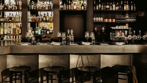Una zona de bar con mostrador, taburetes y estantes llenos de botellas y vasos.