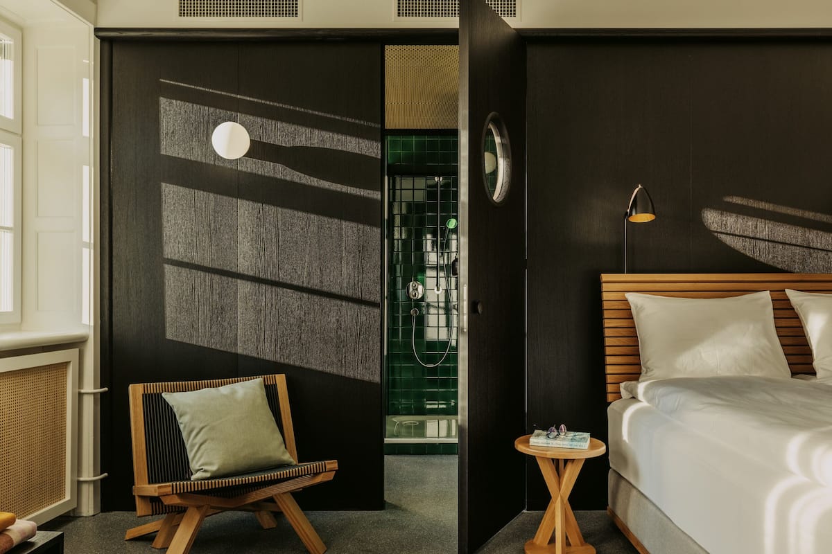 Una habitación de hotel moderna con una cama, una silla de madera, una mesita redonda pequeña y una pared oscura con textura.