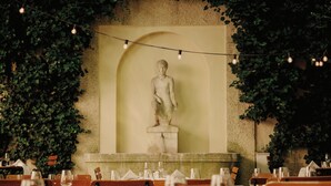Un restaurante con una estatua de piedra al fondo, rodeado de hiedra. Las mesas están preparadas con copas de vino y platos.