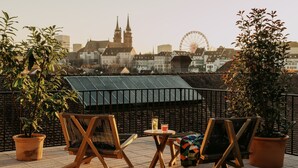 Terraza en la azotea con dos sillas de madera y una mesita pequeña, con vistas a un paisaje urbano que incluye una iglesia y una noria.