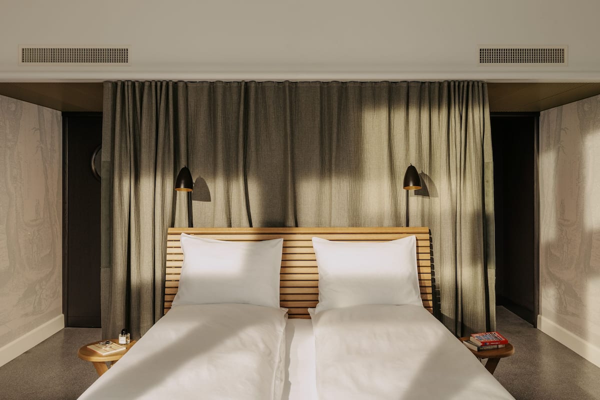 Una cama doble con cabecera de madera, dos mesitas de noche con libros y un gran ventanal con cortinas.