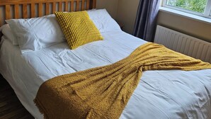 Un lit bien fait, avec du linge de lit blanc et un plaid de couleur moutarde, une tête de lit en bois, une petite étagère avec une plante et une fenêtre avec des rideaux.