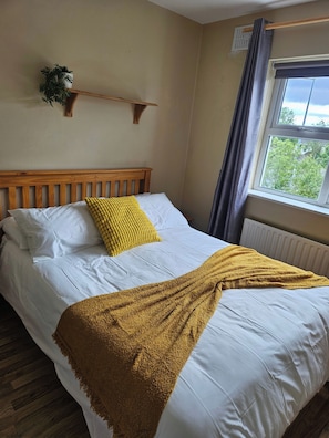 A neatly made bed with white linens and a mustard-colored throw blanket, a wooden headboard, a small shelf with a plant, and a window with curtains. - The Yellow Bollard Galway (Galway)