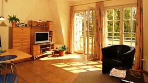 A living room with a TV, wooden furniture, a dining area, and large windows with curtains.