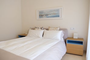 A neatly made bed with white linens, a painting of a harbor scene above it, and a nightstand with a lamp and a power outlet.