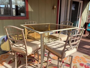 Une table avec un plateau en verre, quatre chaises, une porte avec un heurtoir en laiton et une fenêtre avec des stores.