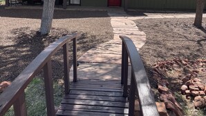 A wooden pathway leading to a building with a red door, surrounded by trees and a covered outdoor seating area. - Spacious centrally located peaceful house Red Rock views and a fenced dog yard! (Sedona)