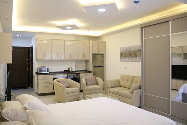 A modern bedroom with a bed, two armchairs, a small kitchenette, and a sliding wardrobe.