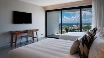 A hotel room with a large bed, a desk, and a TV. There is a balcony with a view of the city and the ocean.