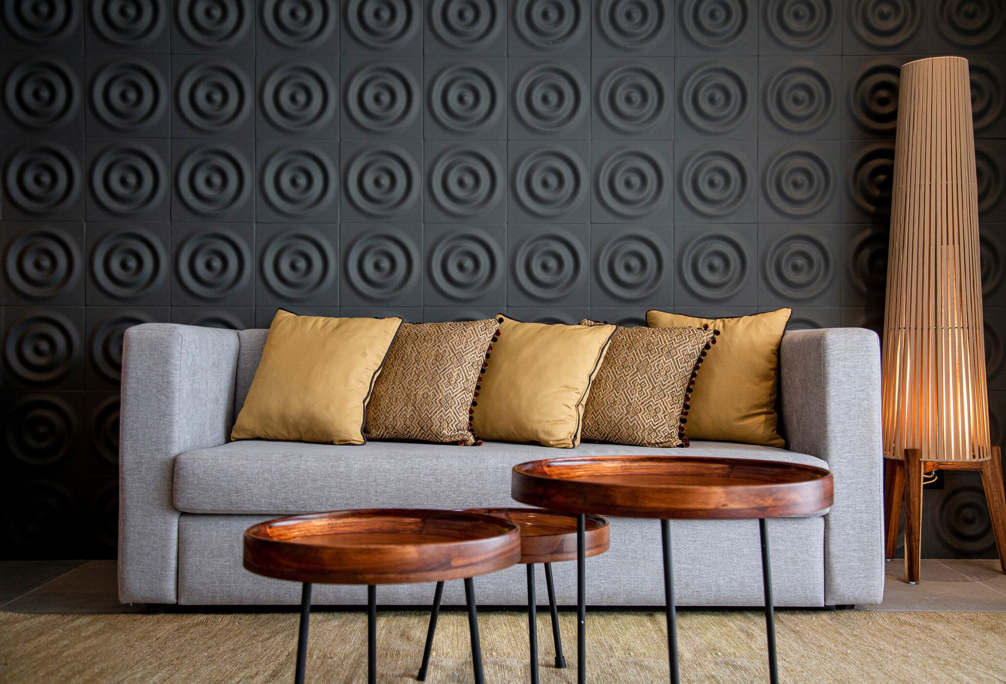 Un sofá gris con cojines estampados, dos mesitas de madera y una lámpara alta y estilizada frente a una pared con textura.