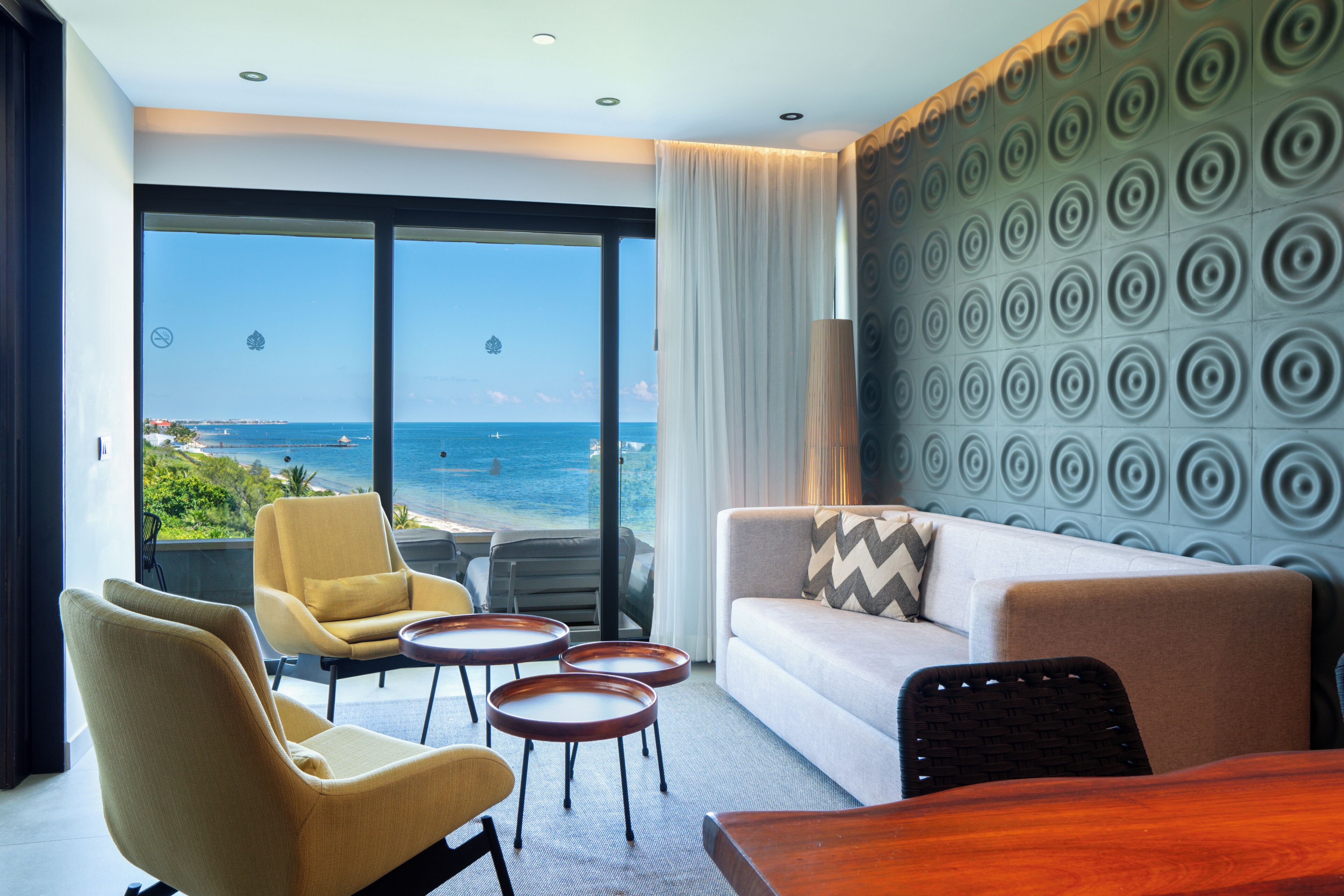 Habitación de hotel moderna con un ventanal grande que ofrece vistas al océano, un sofá, dos sillones, una mesa de madera y un papel tapiz decorado.