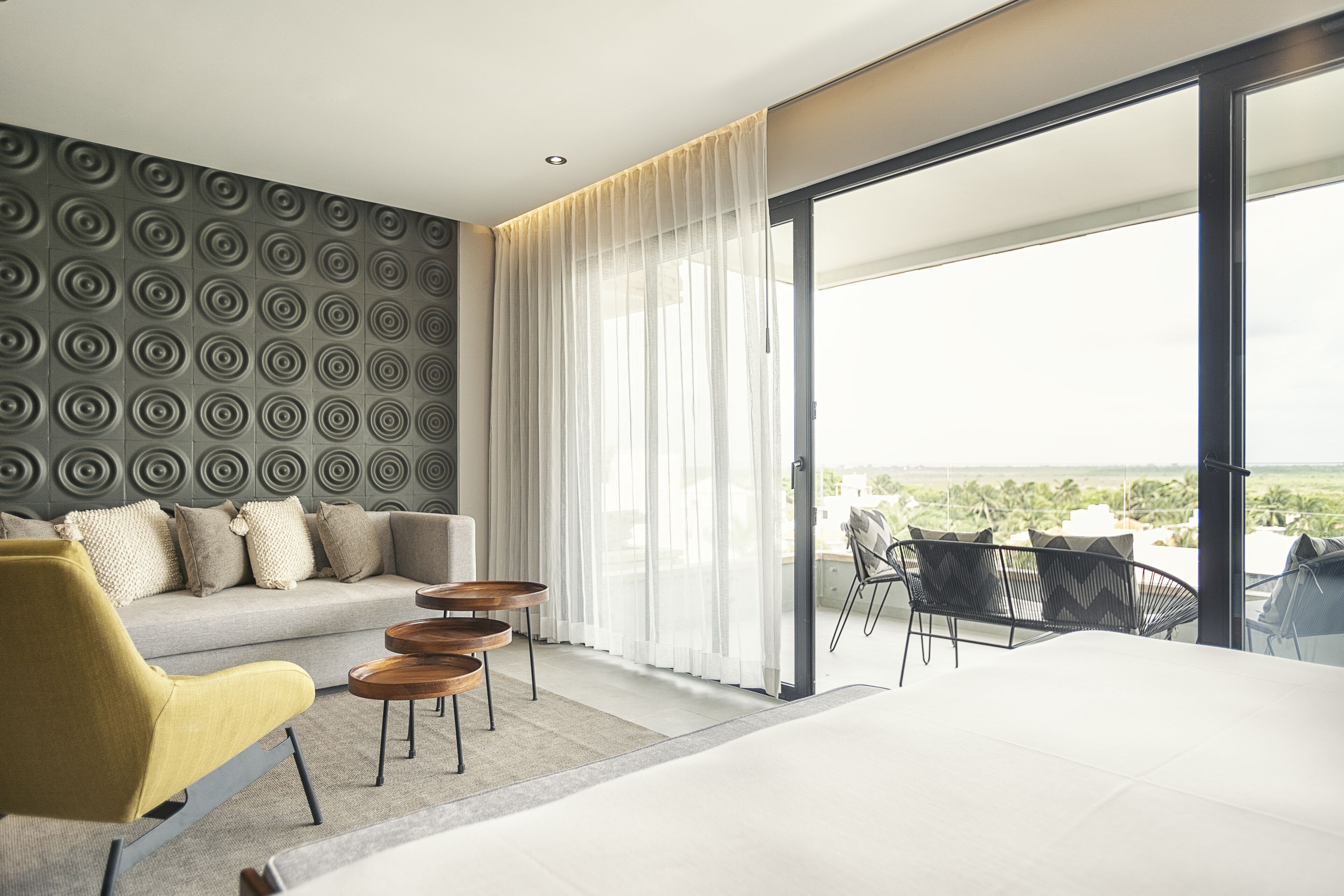 A modern hotel room with a sofa, yellow armchair, and a dining area with a table and chairs.