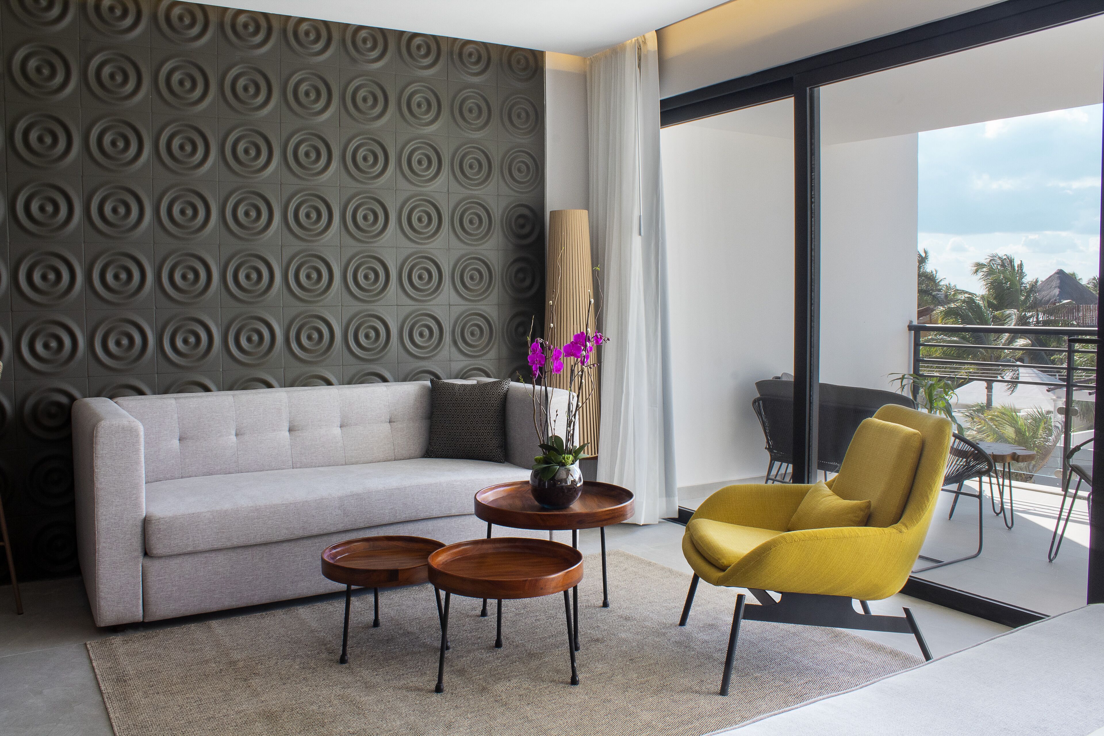 A modern living room with a sofa, two side tables, a yellow armchair, and a view of the balcony.