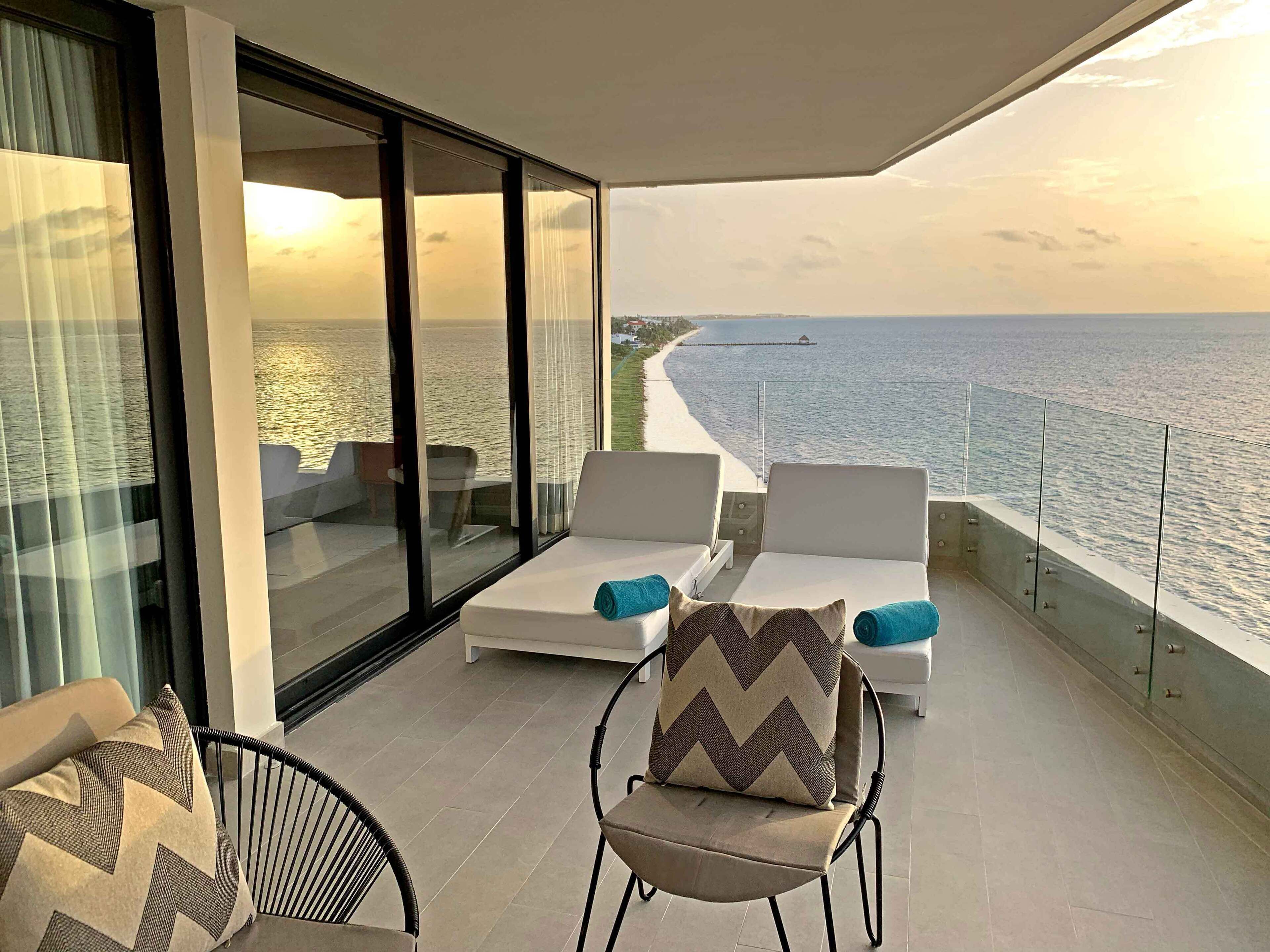 Un balcon avec vue sur la mer, agrémenté de deux fauteuils blancs, dont l’un est orné d’un coussin à motifs et l’autre d’un coussin à rayures en zigzag.