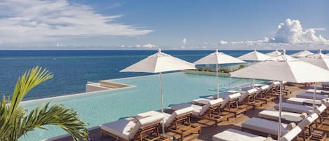 Zona piscina con poltronghe a sdraio e ombrelloni con vista su un mare blu e limpido.