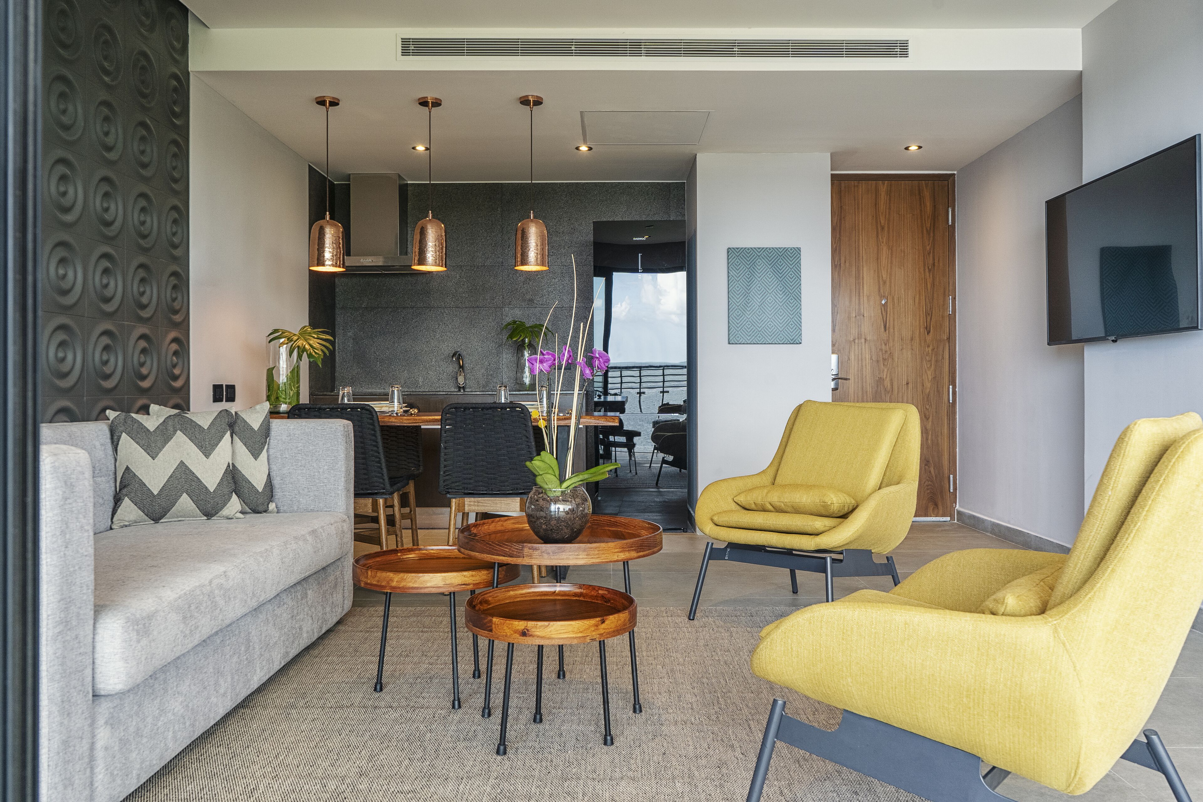 Un salon moderne avec un canapé, un fauteuil jaune et des tables rondes en bois. On aperçoit à l’arrière-plan un coin repas avec une chaise noire et une plante.