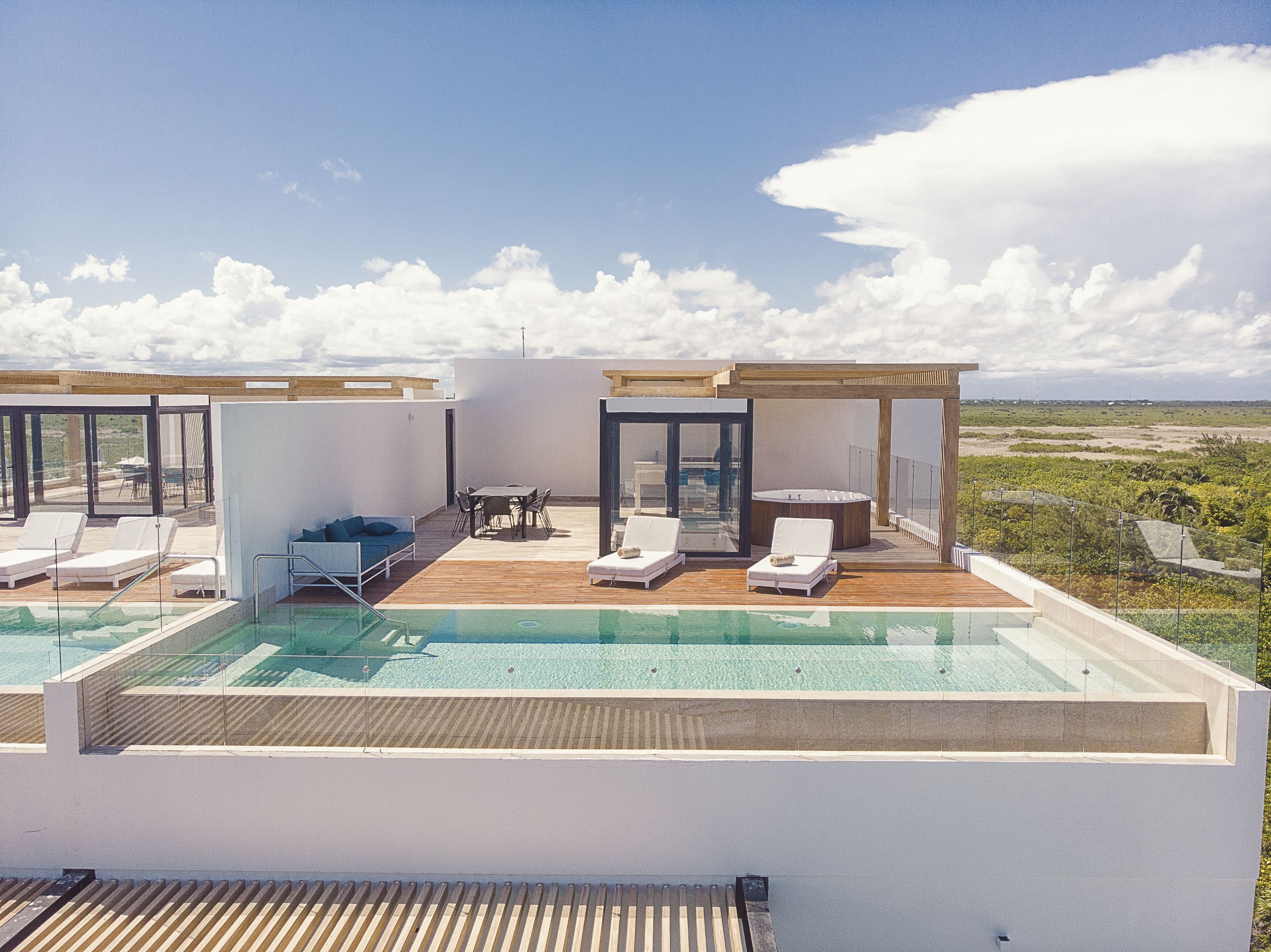 A rooftop pool area with lounge chairs, a hot tub, and a wooden deck overlooking a landscape.