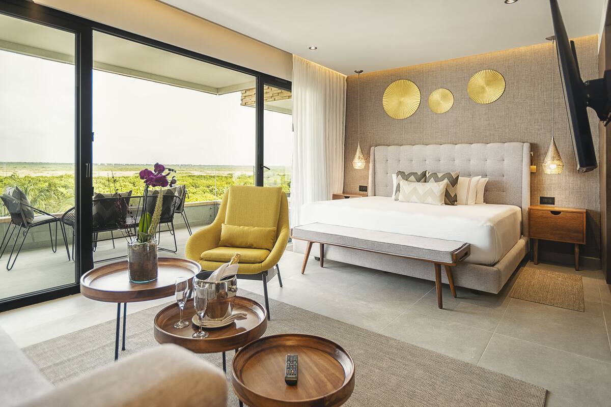 Une chambre d’hôtel moderne avec un grand lit, un fauteuil jaune, une table à manger et une vue sur la plage.