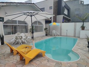 Een zwembad met een terras van stenen tegels, omgeven door witte plastic stoelen en gele ligstoelen.