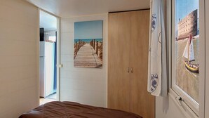 A bedroom with a bed, a wardrobe, a painting of a beach, and a window with a view of a boat. - REDUCTIONS : TO RENT MOBILE HOME IN LA COTINIERE (Saint-Pierre-d'Oléron)