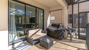 A sunlit patio with a cushioned seating area, a glass table, and a barbecue grill. - 5BR Solterra Disney Retreat w/ Themed Rooms | Tall Ceilings | Game Room | Large pool and spa (Davenport)