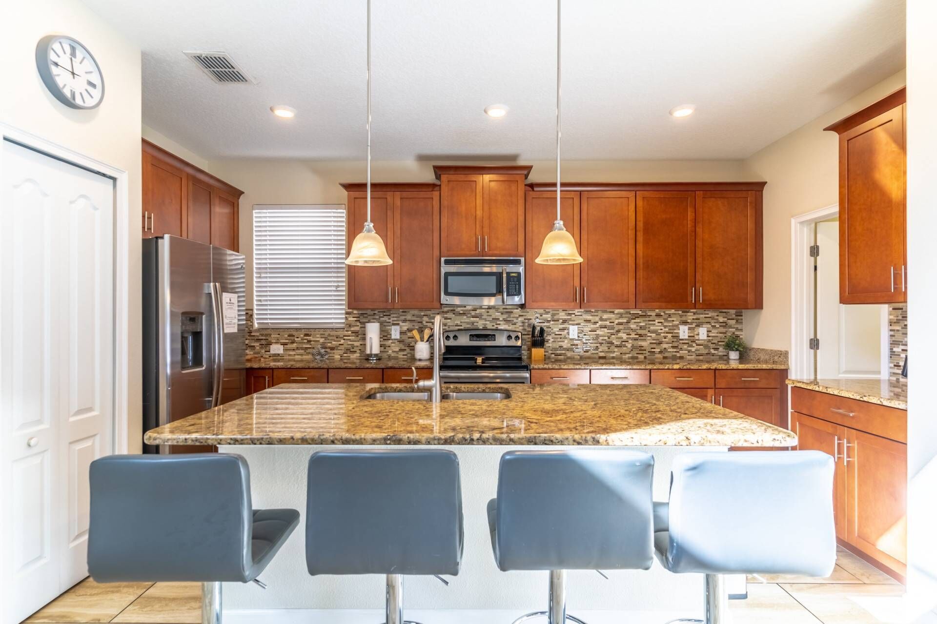 A modern kitchen with a large island, bar stools, wooden cabinets, stainless steel appliances, and pendant lighting.