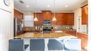 A modern kitchen with a large island, bar stools, wooden cabinets, stainless steel appliances, and pendant lighting. - 5BR Solterra Disney Retreat w/ Themed Rooms | Tall Ceilings | Game Room | Large pool and spa (Davenport)