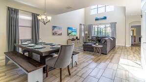 A spacious living area with a dining table set for four, a gray sofa, and a large window providing natural light. - 5BR Solterra Disney Retreat w/ Themed Rooms | Tall Ceilings | Game Room | Large pool and spa (Davenport)