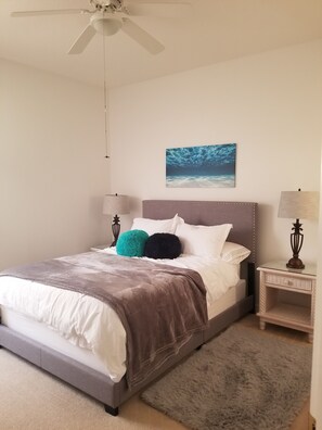 Un lit bien fait, avec une couverture grise et deux oreillers, un ventilateur de plafond, un tableau accroché au mur, deux lampes de chevet et un tapis moelleux.