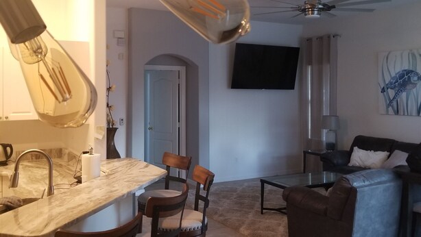 Ett modernt kök med en bänkskiva i marmor, barstolar och hängande lampor. Det finns också en matplats med en soffa och en platt-TV som är monterad på väggen.