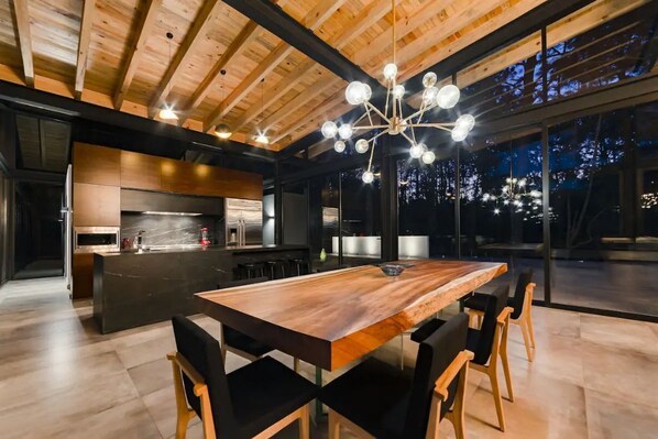 A modern kitchen with a central island, a dining area with a large wooden table, and a view of the night sky through large windows.