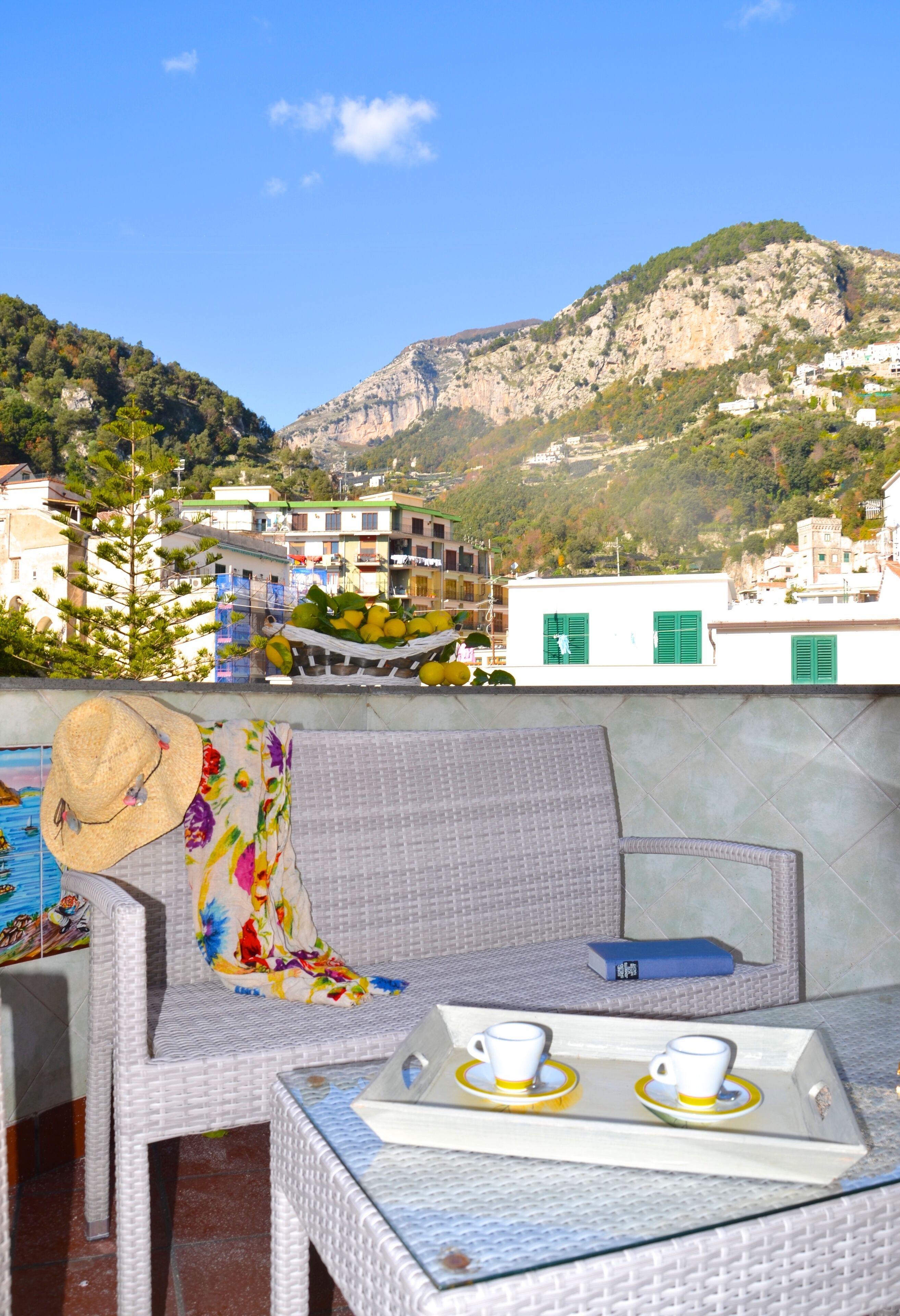 Una terraza con mobiliario de mimbre, una mesa con tazas y platos, y vistas a edificios y montañas.