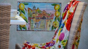 Una silla con funda de estampado floral, una taza blanca con platillo y un cuadro de un edificio con fruta en la pared.