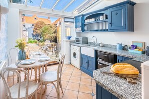 A kitchen with blue cabinets, a microwave, a sink, and a dining area with a table and chairs. - Bracken Cottage, Brancaster Staithe, Norfolk (Brancaster Staithe)