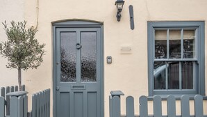 Ein Haus mit grauer Tür, einem Fenster mit Rollläden und einem kleinen Baum davor.