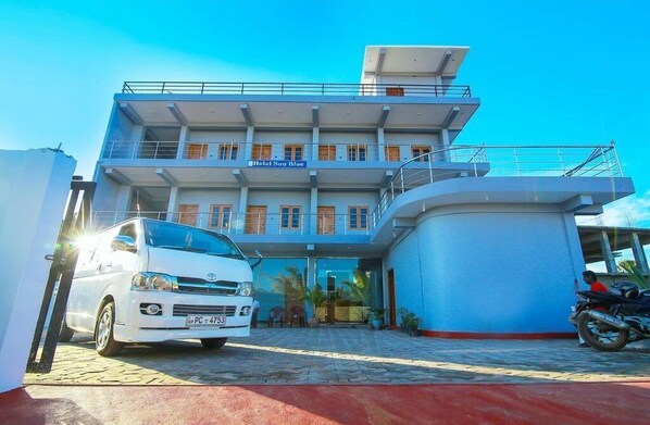 A multi-story hotel with a blue facade, a white car parked in front, and a motorcycle to the right.