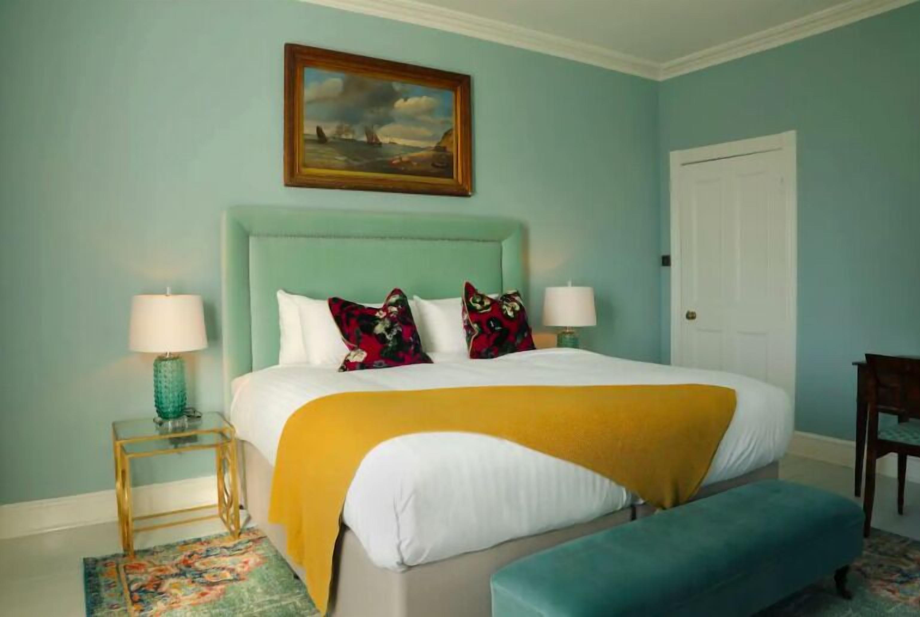 A bed with a yellow throw, a teal upholstered bench, and a painting above it.
