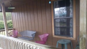 A wooden porch with a white railing, two metal chairs, a small table, and a window with a view of trees. - Whispering Creek Lodging & RV Resort (Newton)