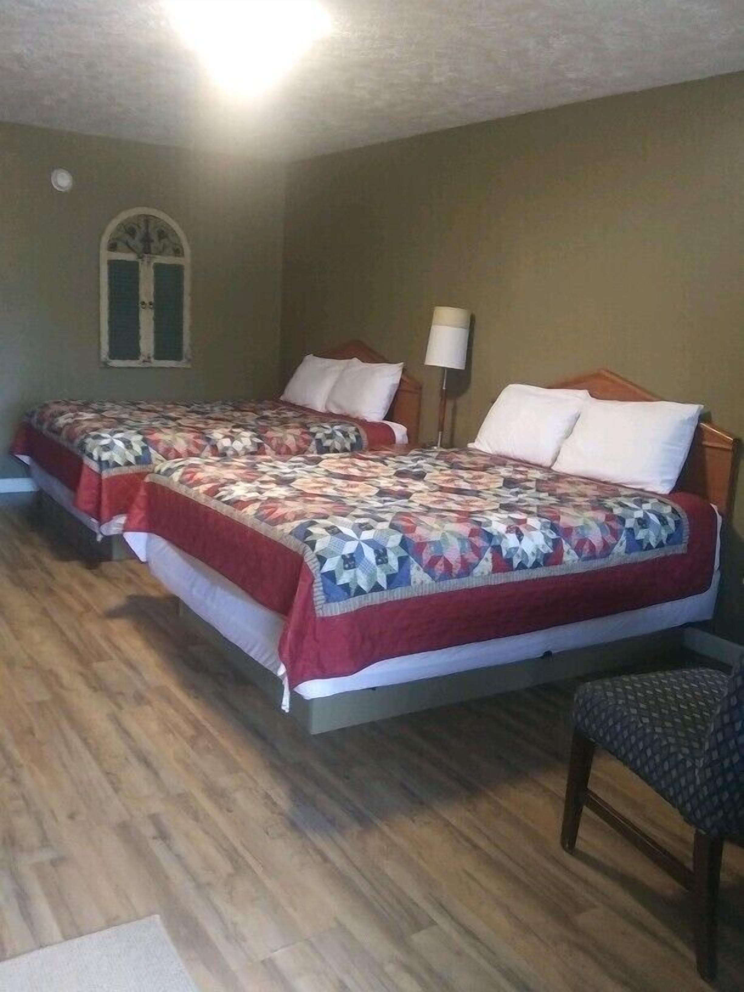 A room with two single beds, each with a patterned bedspread and pillows, a wooden floor, a chair, and a wall-mounted lamp.