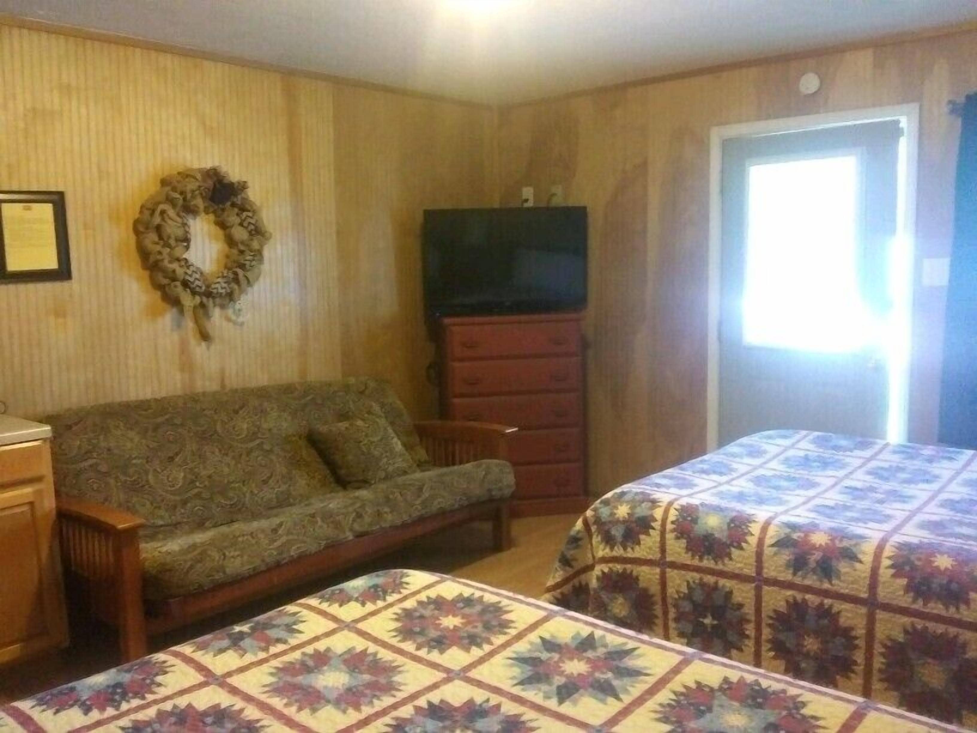A room with a patterned sofa, a dresser, a television, and two beds with patterned quilts.