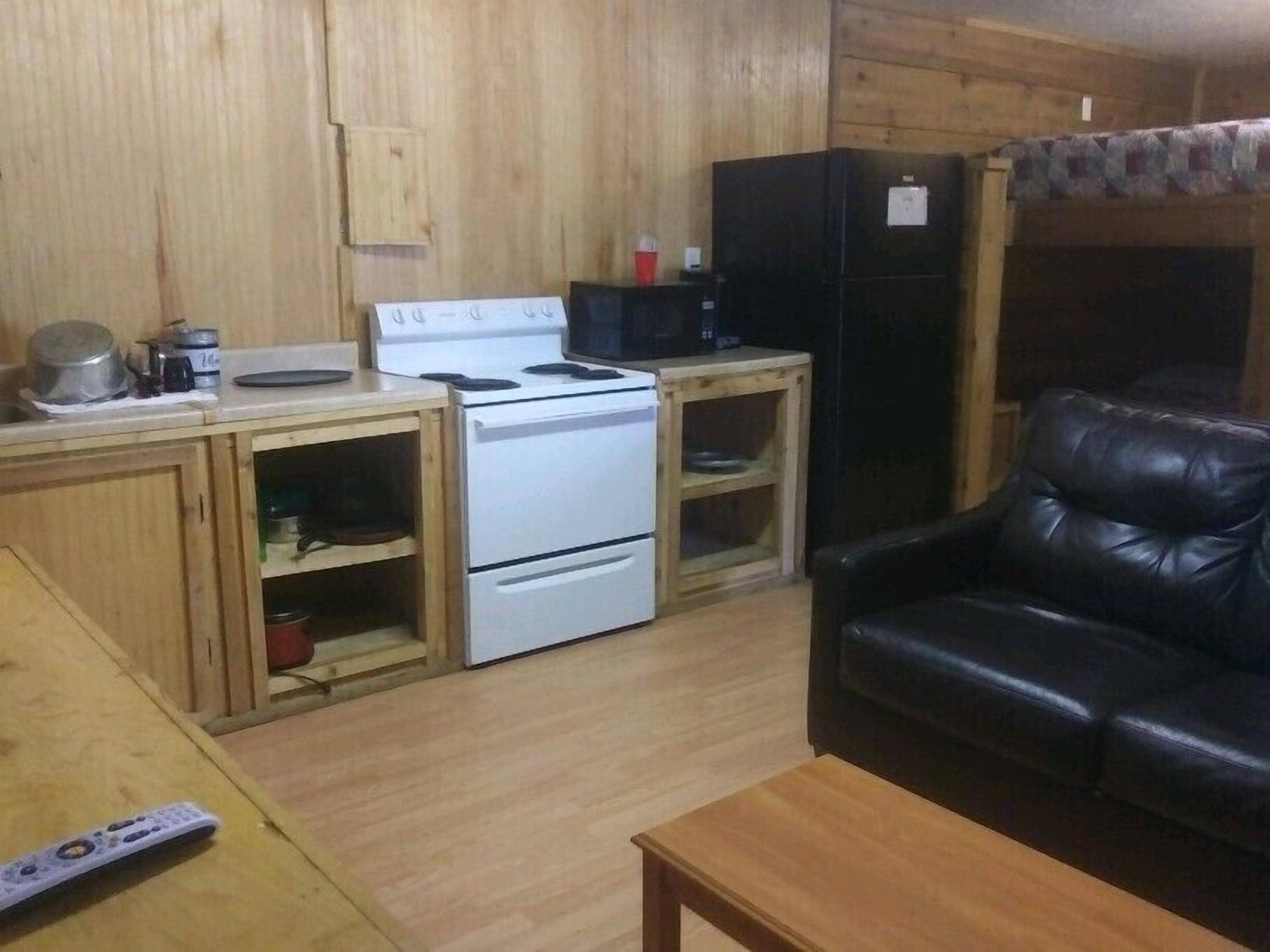A compact kitchen with a stove, refrigerator, and wooden cabinets. A black leather sofa is in the living area.