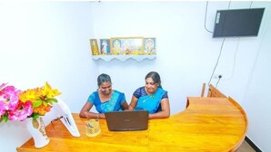 Two individuals seated at a wooden desk with a laptop, a vase of flowers, and a wall-mounted television. - Hotel Sun Blue - Purity in paradise (Alas Garden Trincomale Trincomale)