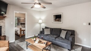 A living room with a gray sofa, a wooden coffee table, a ceiling fan, and a TV mounted on the wall.