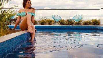 Una persona sentada junto a una piscina con agua azul cristalina, rodeada de plantas y sillas modernas.