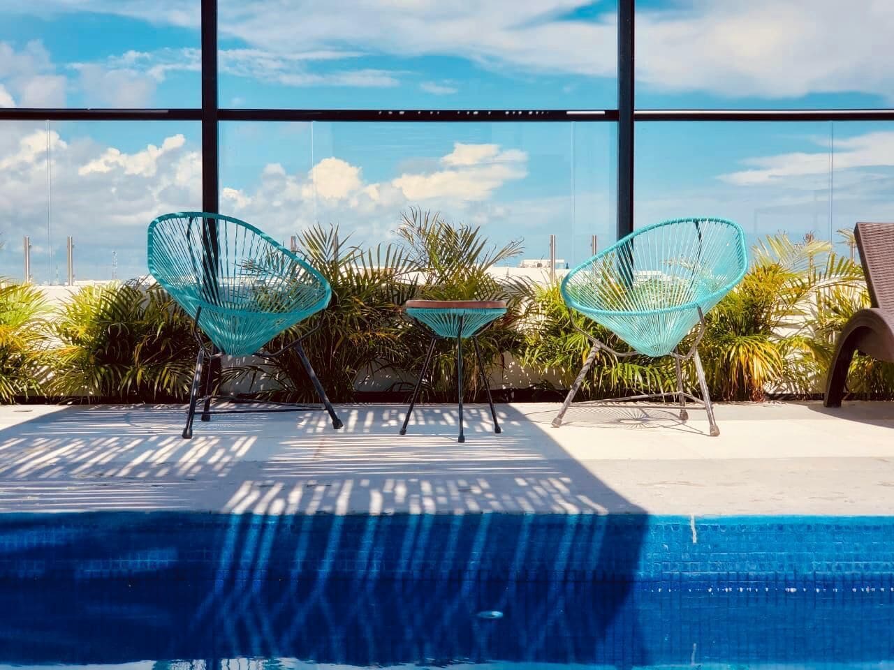 A poolside area with a view of the sky through large windows, surrounded by tropical plants and two turquoise chairs.