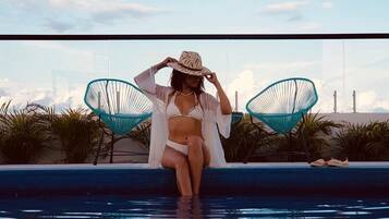 Una persona sentada junto a una piscina, con cielo despejado y palmeras al fondo.